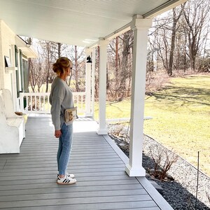 May include: A person wearing a gray sweater, light blue jeans, and white sneakers with black stripes stands on a gray porch. A small beige bag is worn across the body. The porch has white columns and a light blue ceiling.