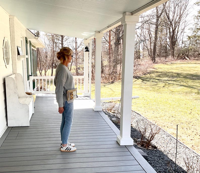May include: A person wearing a gray sweater, light blue jeans, and white sneakers with black stripes stands on a gray porch. A small beige bag is worn across the body. The porch has white columns and a light blue ceiling.