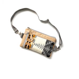May include: A rectangular crossbody bag with a gray adjustable strap. The bag features a beige top and a patterned front with black, white, and orange geometric designs. It has a zipper closure and silver-tone hardware.