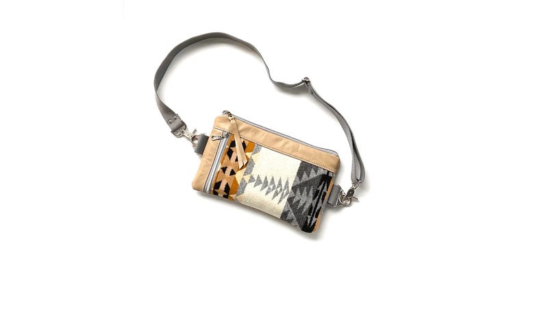 May include: A rectangular crossbody bag with a gray adjustable strap. The bag features a beige top and a patterned front with black, white, and orange geometric designs. It has a zipper closure and silver-tone hardware.