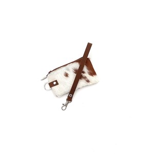 May include: A small, rectangular cowhide pouch with a zipper closure and a brown leather strap. The pouch features a white background with brown spots. A silver-colored key ring and clasp are attached.