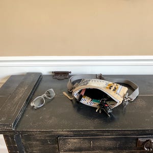 May include: A black wooden table with a distressed finish holds a beige fanny pack with a geometric pattern, sunglasses, and keys. The fanny pack is open, revealing its contents. The background is a neutral wall.