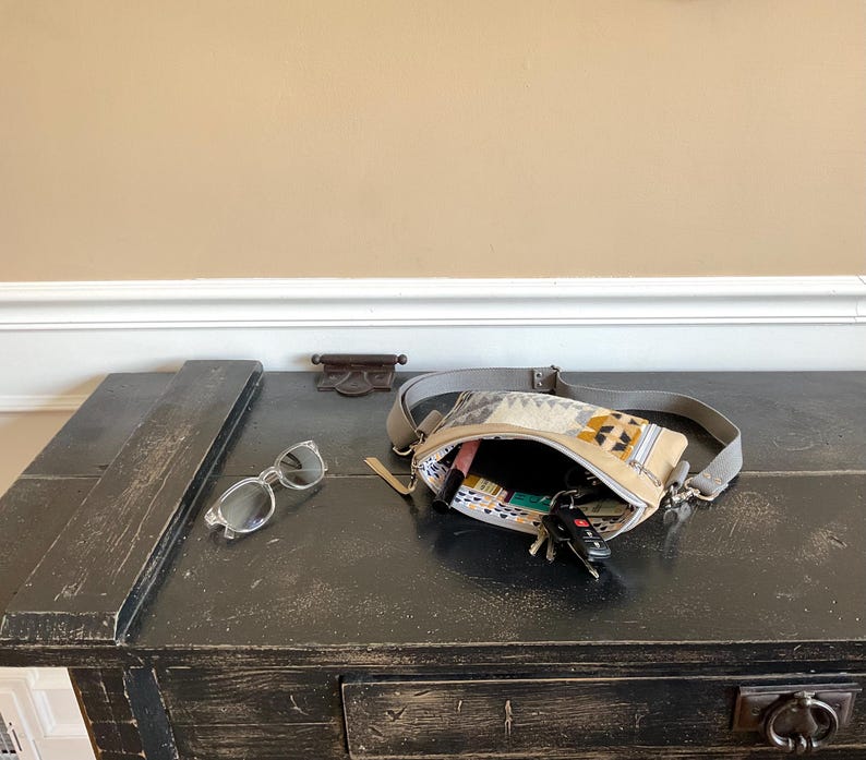 May include: A black wooden table with a distressed finish holds a beige fanny pack with a geometric pattern, sunglasses, and keys. The fanny pack is open, revealing its contents. The background is a neutral wall.
