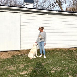 May include: A woman in a gray sweatshirt and blue jeans stands next to a light-colored Labrador Retriever dog on a grassy lawn. The dog is wearing a black collar. The background features a white building with a black roof and bare trees.