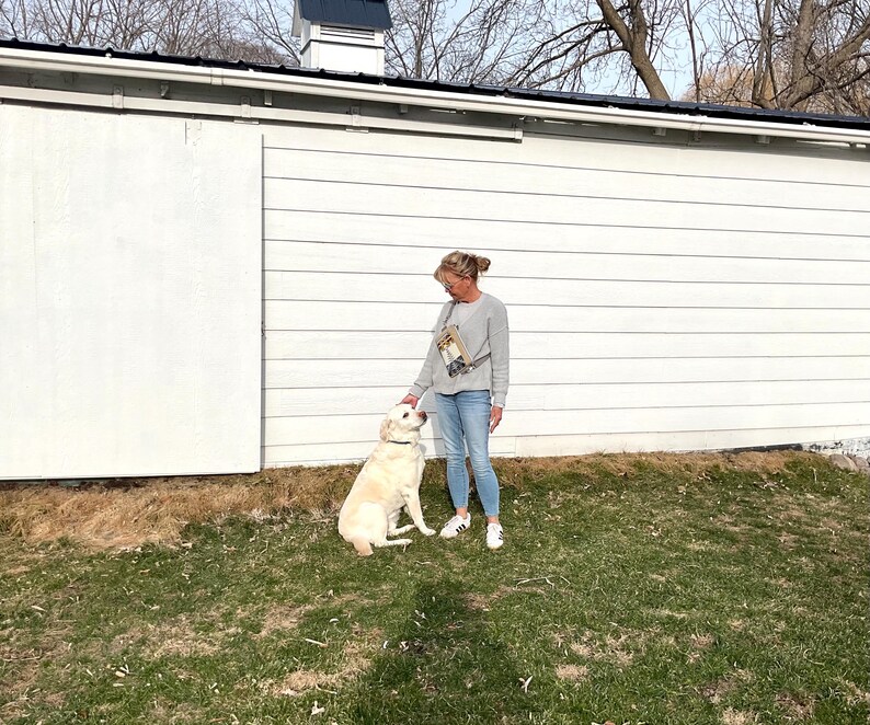 May include: A woman in a gray sweatshirt and blue jeans stands next to a light-colored Labrador Retriever dog on a grassy lawn. The dog is wearing a black collar. The background features a white building with a black roof and bare trees.