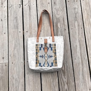May include: A white canvas tote bag with brown leather handles and a decorative front panel. The panel features a geometric pattern in shades of blue, tan, and gray. The bag is secured with a metal clasp.