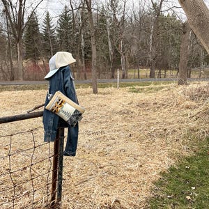 May include: A denim jacket and jeans hang on a metal fence post, with a beige baseball cap perched on top. A patterned clutch with geometric designs is attached to the jacket. The background features a field and trees.