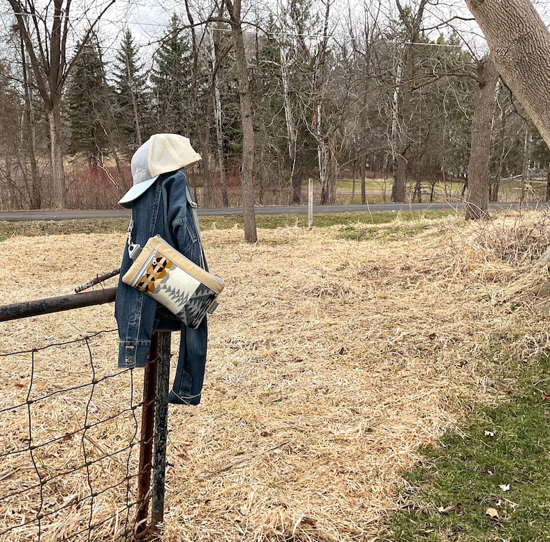 May include: A denim jacket and jeans hang on a metal fence post, with a beige baseball cap perched on top. A patterned clutch with geometric designs is attached to the jacket. The background features a field and trees.