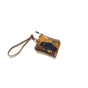 May include: A small, rectangular coin purse with a brown leather wrist strap. The purse features a tapestry design with a dark blue bison silhouette on a tan and brown background. A small zipper and metal hardware complete the design.