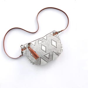 May include: A gray and white patterned crossbody bag with a brown leather strap and a matching tassel. The bag features a geometric diamond design and is set against a white background.