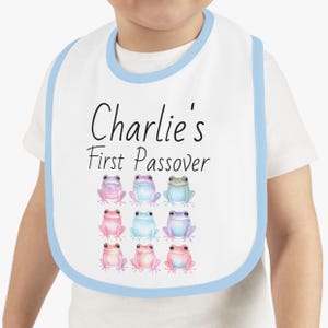 May include: A white bib with a light blue trim, featuring the text "Charlie's First Passover" and a row of nine colourful frog illustrations.