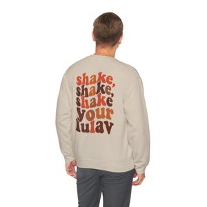 Puede incluir: Sudadera beige con la frase "shake, shake, shake your lulav" impresa en la espalda en tonos naranja, marrón y rojo. La sudadera es de color neutro y tiene cuello redondo.