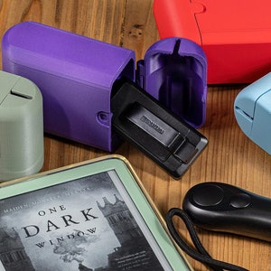May include: A collection of colorful 3D-printed cases and accessories. The image features a purple case with a black object inside, along with green, red, and blue cases. An e-reader with the book "One Dark Window" is also visible, along with a black remote.