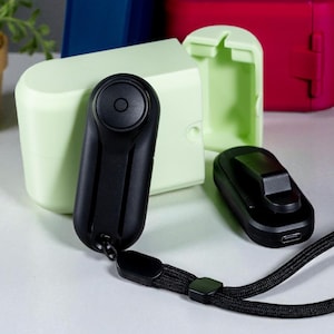 May include: A collection of small, black electronic devices with a wrist strap, alongside colorful rectangular cases in blue, red, and green. A small potted plant and a silver alarm clock are also present on a white surface.