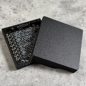 Może przedstawiać: Czarny elektroniczny syntezator muzyczny z siatką przycisków i pokręteł. Syntezator jest otwarty, a pokrywa znajduje się obok. Pokrywa jest również czarna i ma fakturowaną powierzchnię.