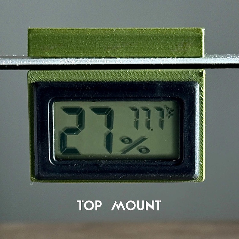 May include: A green digital hygrometer with a black display showing a temperature of 27 degrees and a humidity of 11.1%. The hygrometer is mounted on a surface with the text "TOP MOUNT" below it.