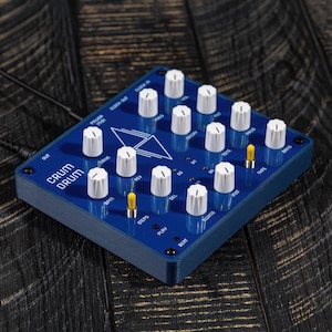 May include: A blue, compact drum machine with white knobs and a yellow button. The machine has labels for "CRUM DRUM", "BPM", "STEPS", "DEL", "PLAY", "BEAT", "SWING", "INST", "TYPE", "RING", "TONL", "DECIN", "50", "1", and "POWER".