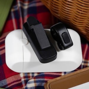 Puede incluir: Una funda de carga blanca con dos dispositivos electrónicos negros encima. Un dispositivo tiene un clip y el otro tiene una forma pequeña y redondeada. Los artículos descansan sobre una superficie a cuadros roja, blanca y azul, con una cesta de mimbre en el fondo.