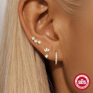 May include: A close-up of an ear displaying three gold earrings. One is a curved bar with star-shaped gems, another a floral drop, and the third a small hoop with gems. The earrings are set against the backdrop of blonde hair.