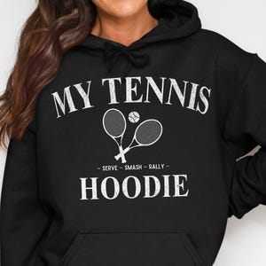 Op de afbeelding: Zwarte hoodie met witte tekst "MY TENNIS HOODIE". Het ontwerp bevat twee gekruiste tennisrackets en een tennisbal. Onder de rackets staat de tekst "SERVE - SMASH - RALLY".