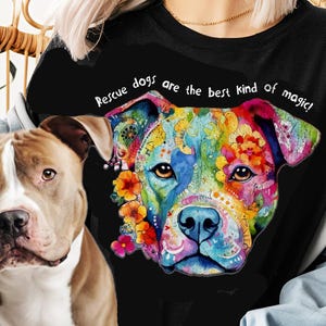以下が含まれることがあります： カラフルな犬の顔のイラストと「レスキュー犬は最高の魔法です！」というテキストが書かれた黒いTシャツ。茶色と白のピットブルテリアが、シャツを着ている人の前に座っています。