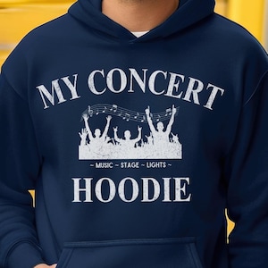 Op de afbeelding: Marineblauwe hoodie met een voorzak en witte tekst "MY CONCERT HOODIE". Het ontwerp bevat een afbeelding van silhouetten van mensen met hun armen omhoog en de woorden "MUSIC - STAGE - LIGHTS -" eronder.