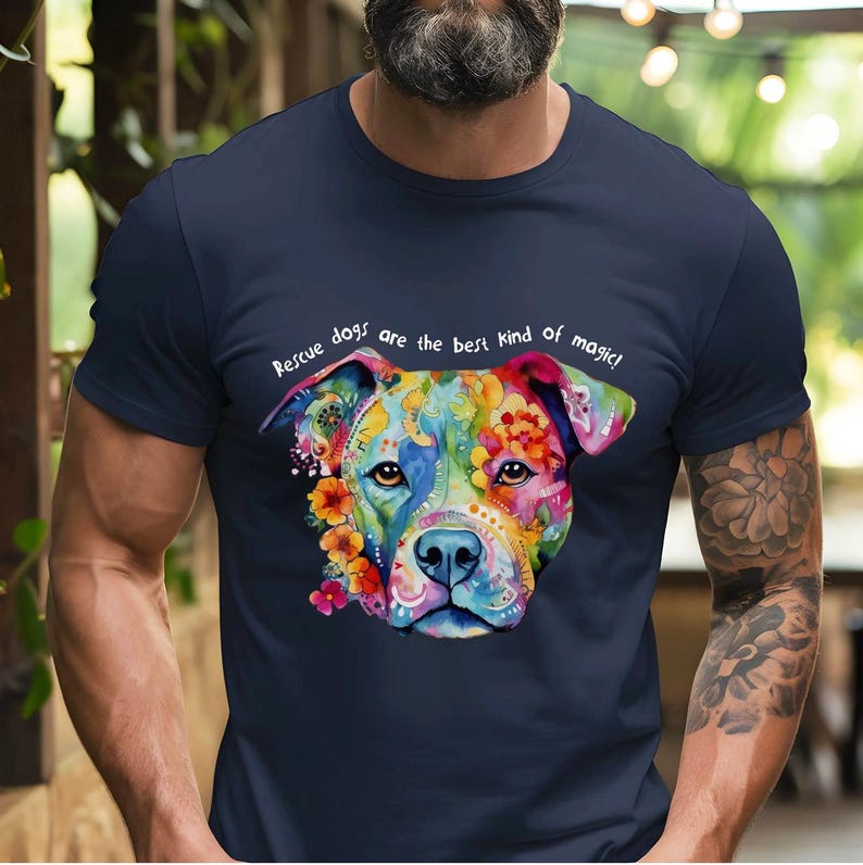 以下が含まれることがあります： カラフルな犬の顔のイラストが描かれたネイビーブルーのTシャツ。Tシャツには「レスキュー犬は最高の魔法です！」という文字がプリントされています。