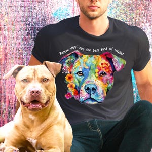 以下が含まれることがあります： 黒いTシャツを着た人が、犬のカラフルなイラストと「レスキュー犬は最高の魔法です！」という文字をプリントしています。茶色と黄褐色のピットブルテリアが前景に座っています。