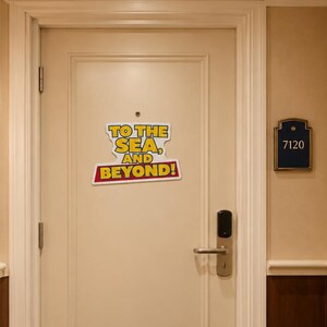 May include: A cream-coloured door featuring a sign with yellow and red text that reads TO THE SEA, AND BEYOND! The door includes a silver handle, a digital lock, and a peephole. To the right, a dark blue plaque displays the number 7120.