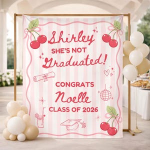 Può includere: Un banner a righe rosa e bianche con il testo "Shirley She's Not Graduated!" e "Congrats Noelle Class of 2026". Il banner è decorato con ciliegie, un diploma e altre grafiche celebrative. Ci sono palloncini su entrambi i lati.