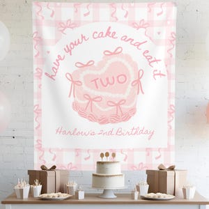 May include: Pink and white gingham backdrop with the text "Have your cake and eat it two" and "Harlow's 2nd Birthday". A pink and white cake with a heart-shaped top is in front of the backdrop. There are balloons and other decorations on a table in front of the backdrop.