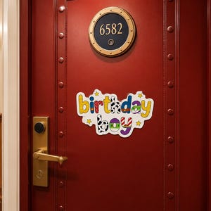 May include: A decorative sign with the text birthday boy in colourful, patterned letters attached to a dark red door. The sign features various designs including cow print, polka dots, and stripes, accented by small yellow stars on a white background.