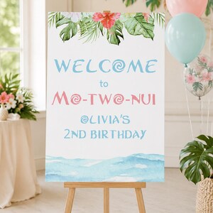 K&ouml;nnte beinhalten: Ein wei&szlig;es Willkommensschild auf einer Holzstaffelei, verziert mit tropischen Blumen und Bl&auml;ttern. Auf dem Schild steht "WELCOME to Mo-TWO-NUI @OLIVIA'S 2ND BIRTHDAY" in blauer und rosa Schrift. Ballons in Rosa, Blau und transparent mit Blumendesigns sind im Hintergrund.