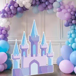 Puede incluir: Un arco de globos de color morado, azul y blanco con un recorte de castillo de cartón en frente. El castillo está decorado con detalles azul claro y morado.