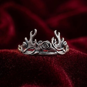May include: A silver-toned ring with an antler and rose design. The ring features two antler-like structures on either side of a central rose, with additional rose details. The ring is set against a deep red velvet background.