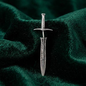 May include: A detailed, silver-toned miniature sword pin. The pin features a textured handle, a crossguard, and an engraved blade. The pin is set against a dark green, velvet-like background, creating a striking contrast. The design is reminiscent of fantasy or historical weaponry.