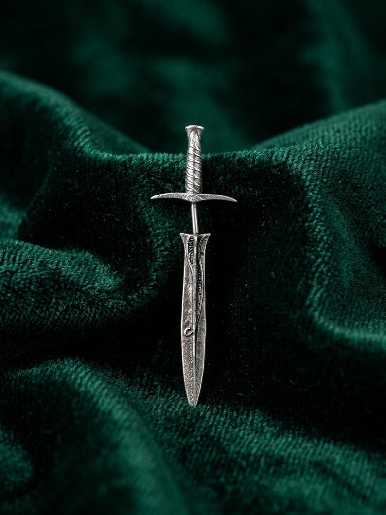 May include: A detailed, silver-toned miniature sword pin. The pin features a textured handle, a crossguard, and an engraved blade. The pin is set against a dark green, velvet-like background, creating a striking contrast. The design is reminiscent of fantasy or historical weaponry.