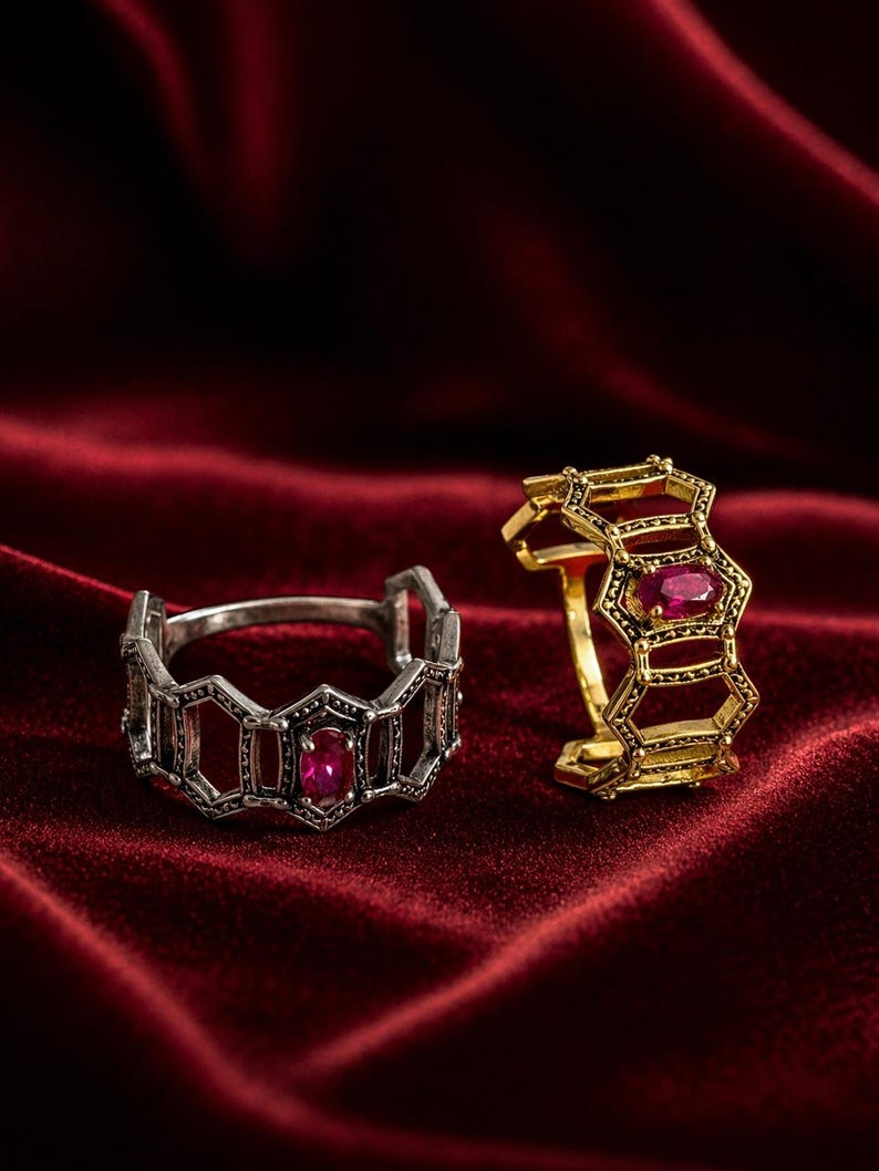 May include: Two rings with geometric designs and ruby gemstones. One ring is silver-toned, and the other is gold-toned. The rings are displayed on a deep red velvet surface.
