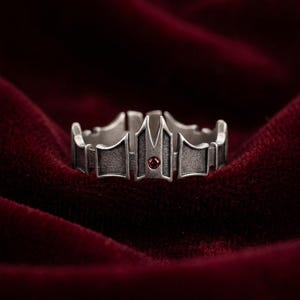 Könnte beinhalten: Ein silberfarbener Ring mit Kronenmuster, der einen kleinen, runden, roten Edelstein in der Mitte aufweist. Der Ring hat eine detaillierte, strukturierte Oberfläche und ist vor tiefrotem Samt drapiert.