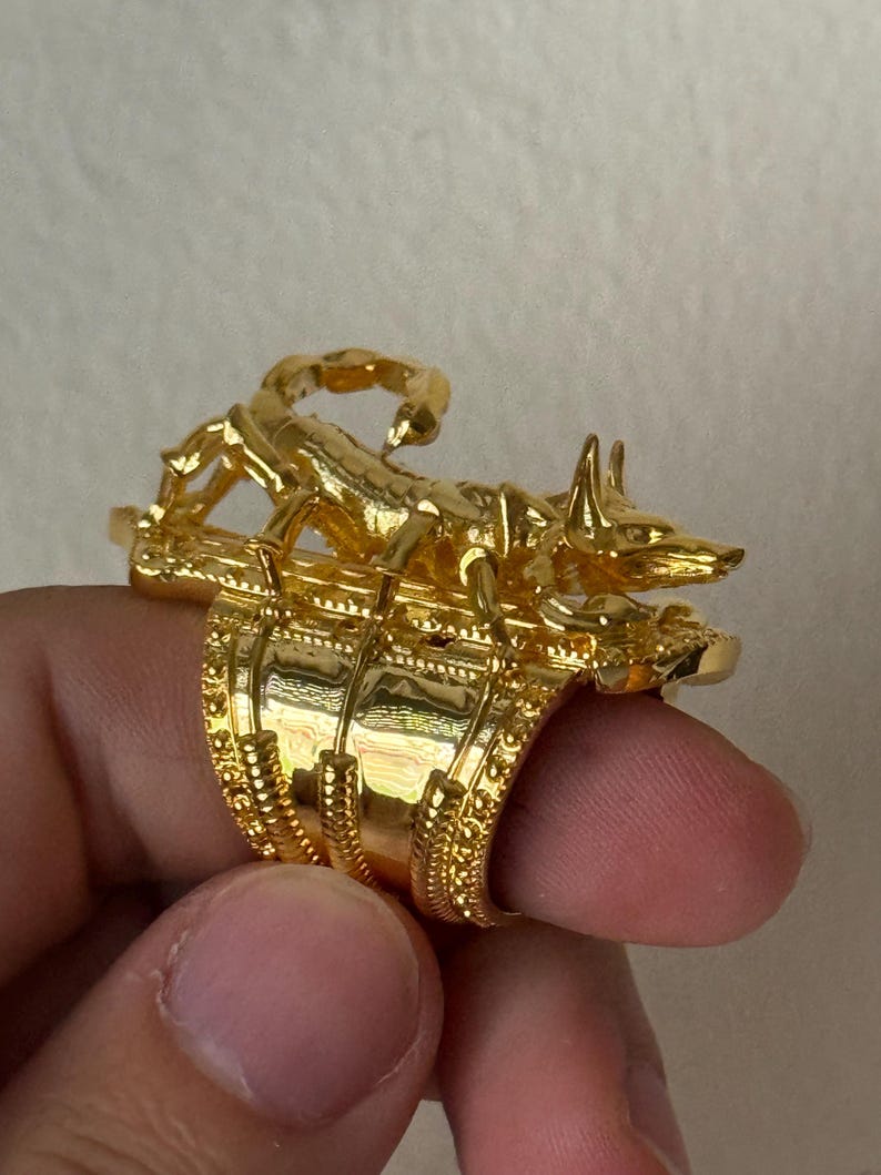 Puede incluir: Un anillo de oro con un dise&ntilde;o detallado que presenta un escorpi&oacute;n y un chacal. El anillo se lleva en un dedo.