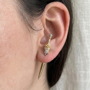 May include: A close-up of an ear with two earrings. The first earring is a gold and silver stud with a decorative top. The second earring is a long, gold, pointed spike that extends from the back of the ear.