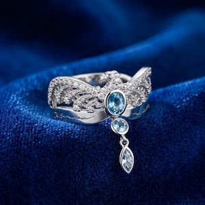 Puede incluir: Un anillo plateado con un diseño de pájaro, con una gran gema azul ovalada y gemas azules más pequeñas. El anillo tiene detalles intrincados y una gema colgante en forma de lágrima. El anillo está sobre un fondo azul oscuro.