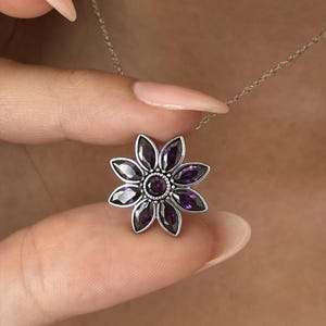 May include: A silver-toned necklace featuring a flower-shaped pendant. The pendant has a central round purple stone surrounded by eight marquise-cut purple stones arranged like petals, all set within a detailed silver metal frame on a thin chain.