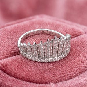Puede incluir: Un anillo plateado con diseño de corona, incrustado con pequeñas piedras brillantes. El anillo se encuentra sobre una superficie de terciopelo rosa. La inscripción "TOGETHER IN PARIS" está grabada en el interior.