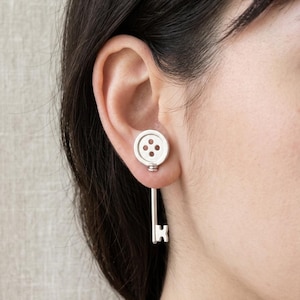 May include: Silver-toned button and key earrings. The button-shaped stud features three small holes, while the key design hangs below. The earrings are worn on the ear, showcasing a unique and playful jewelry design.