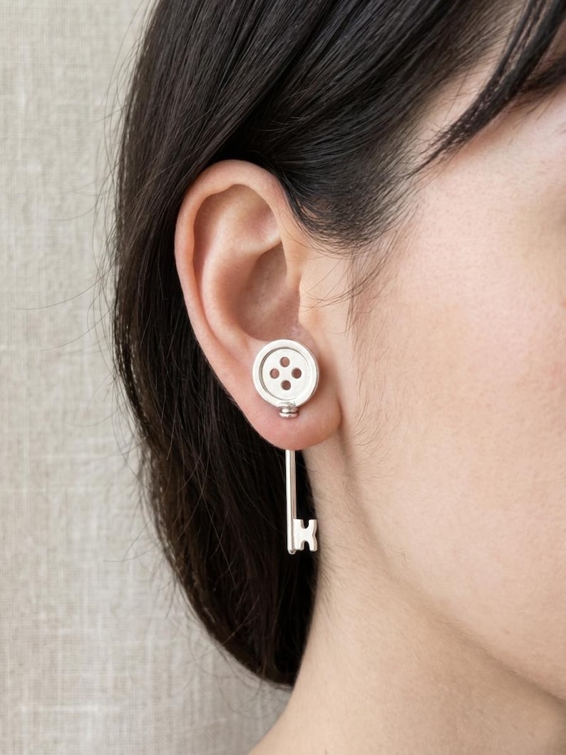 May include: Silver-toned button and key earrings. The button-shaped stud features three small holes, while the key design hangs below. The earrings are worn on the ear, showcasing a unique and playful jewelry design.
