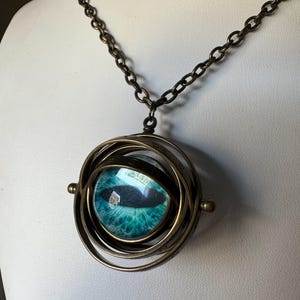Peut inclure: Un collier en laiton avec un pendentif tournant représentant un œil bleu avec une pupille noire. Le pendentif est entouré de plusieurs anneaux.
