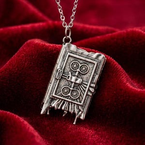 May include: Silver-toned pendant necklace featuring a detailed, open book design. The pendant has intricate embossed patterns and a small clasp. The necklace hangs on a delicate chain against a deep red velvet background.