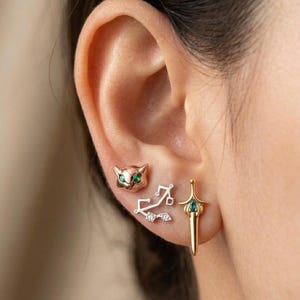 May include: A collection of three earrings: a cat face with green eyes, a Sagittarius symbol, and a sword with a green gem. The earrings are gold and silver in color and are displayed on an ear.