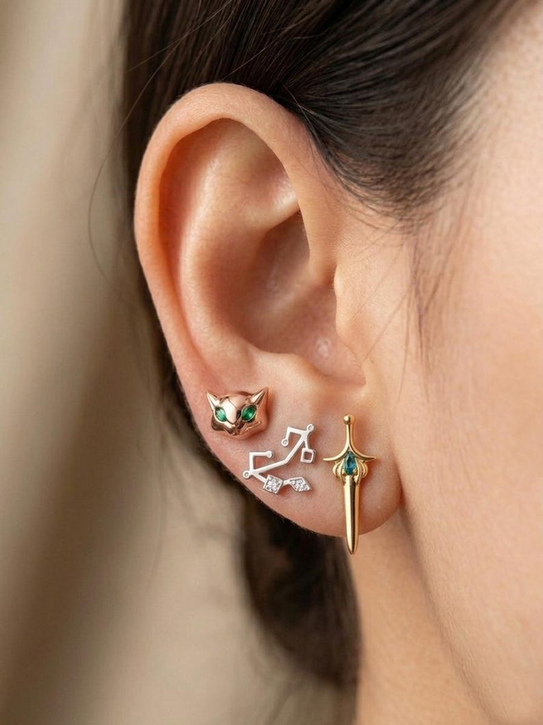 May include: A collection of three earrings: a cat face with green eyes, a Sagittarius symbol, and a sword with a green gem. The earrings are gold and silver in color and are displayed on an ear.